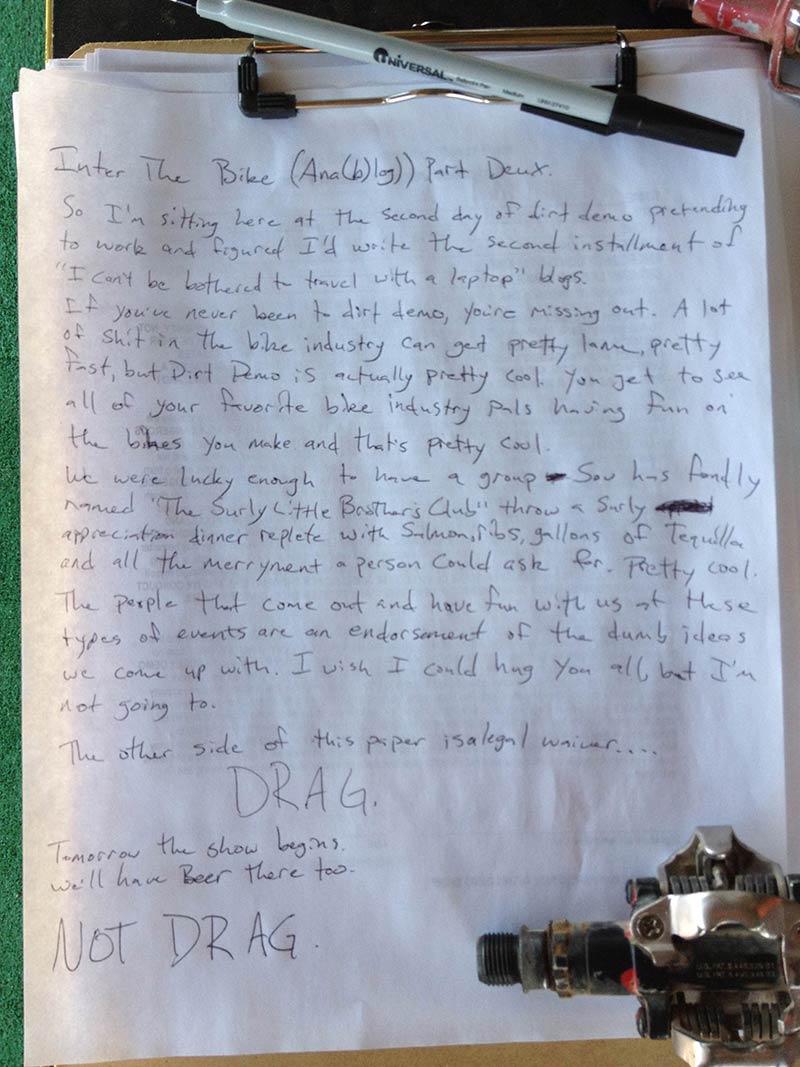 Downward view of a letter, written on a sheet of paper, attached to a clipboard with a pen up top and bike pedal below