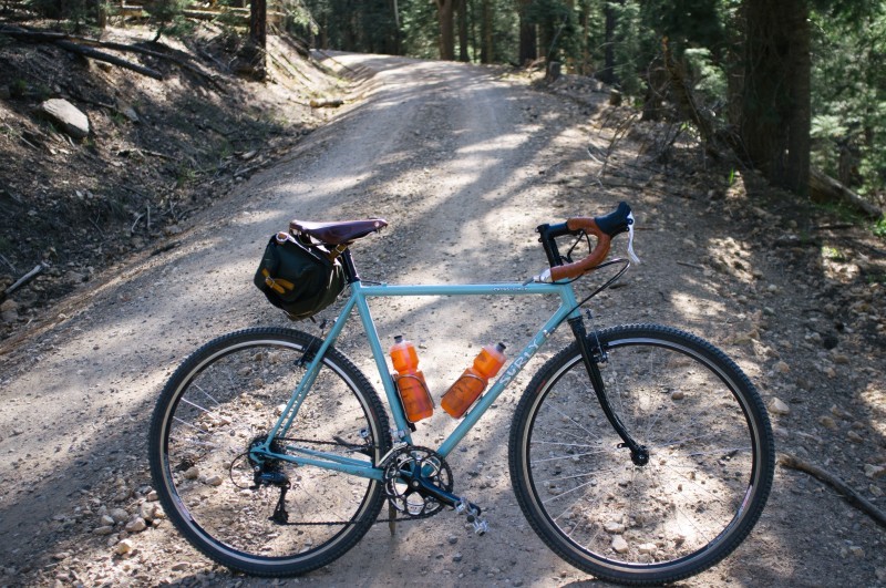 Right side view of a light blue Surly Cross Check bike, parked across a gravel road in the forest