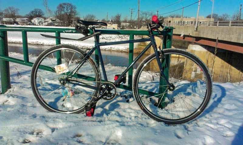 Right side view of a Surly bike, parked on snow against a green gate, on top of a high river bank, with bridge behind