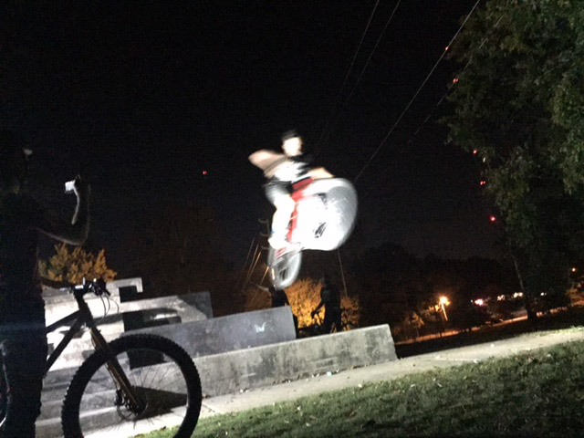 Blurry front view of a cyclists jumping their bike off of a cement step, at night
