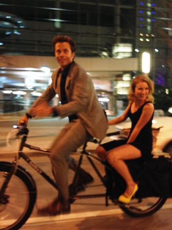 Left side view of a Surly Big Dummy bike, with a cyclist in a suit and a person in a dress on back, at night on a street
