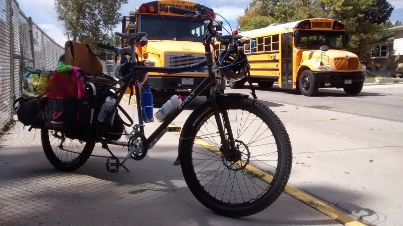 Right side view of a black Surly Big Dummy bike with gear, parked across a sidewalk with school buses in the background