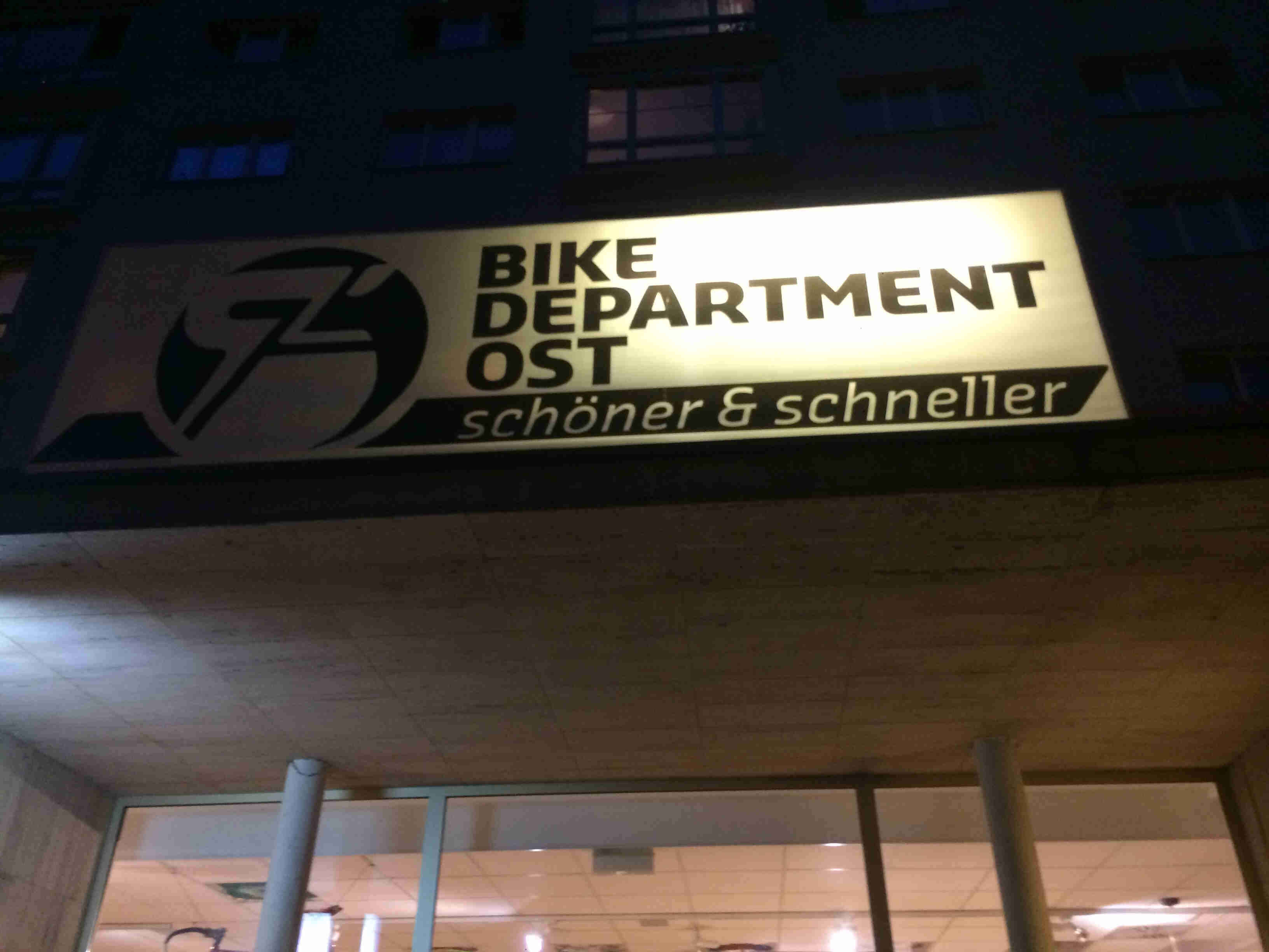 A lit up sign showing, Bike Department OST - schoner & schneller, above the glass front doors, on a building at night