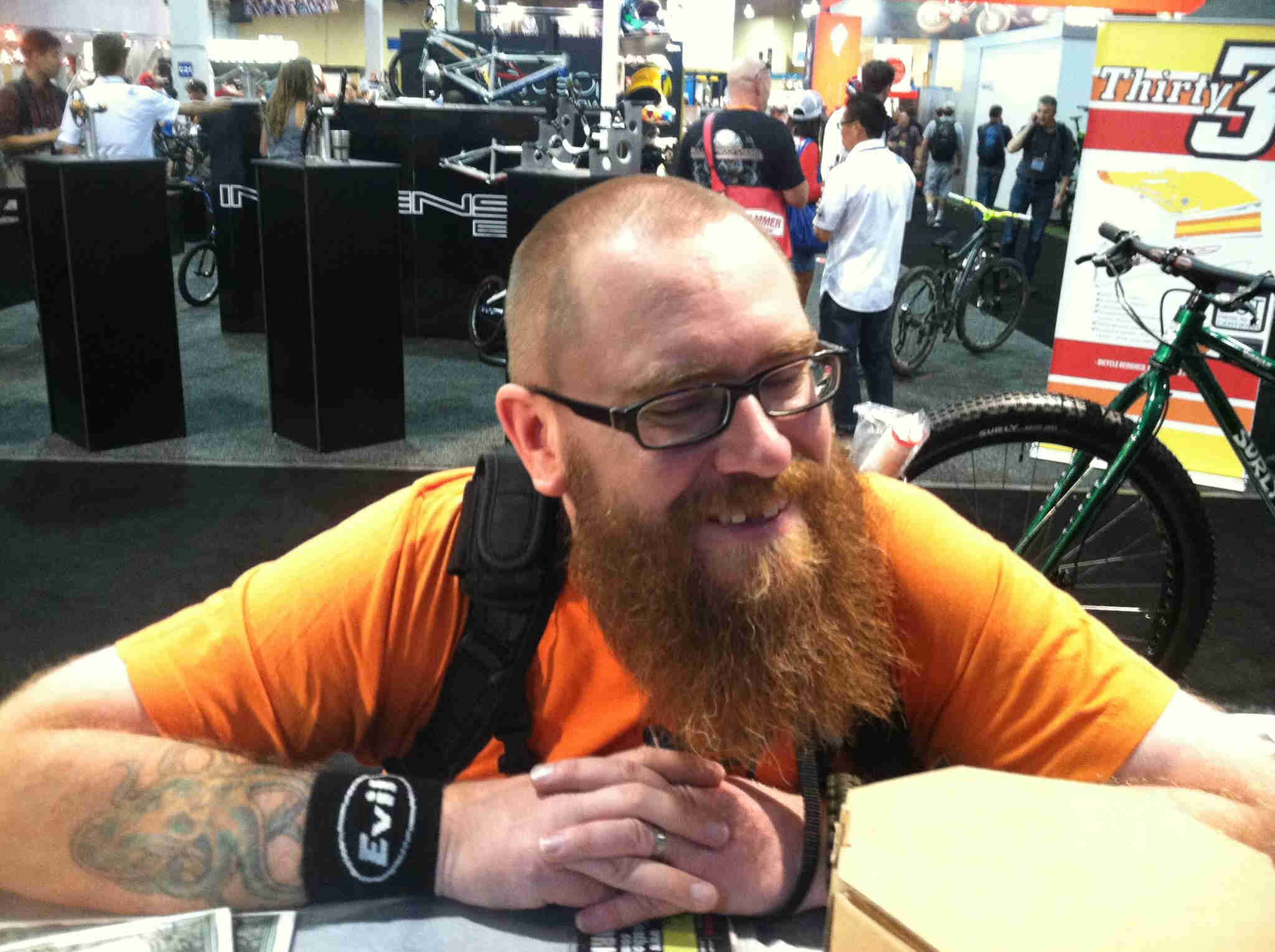 Front, chest up view of a person with a beard, leaning over a table, with tradeshow displays behind him
