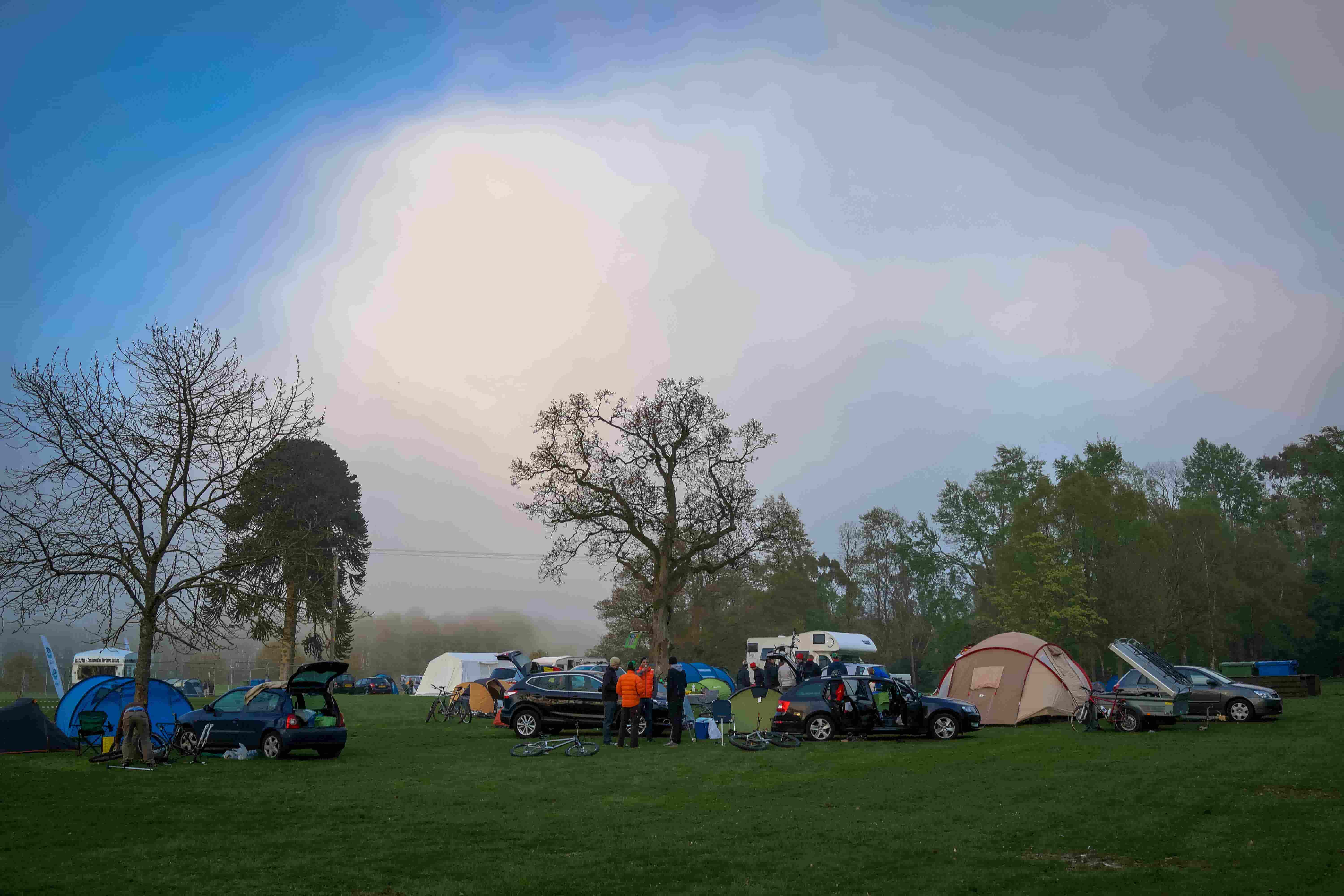A campsite, with people, tents and cars, in a grass field with trees behind them, and a hazy sun above