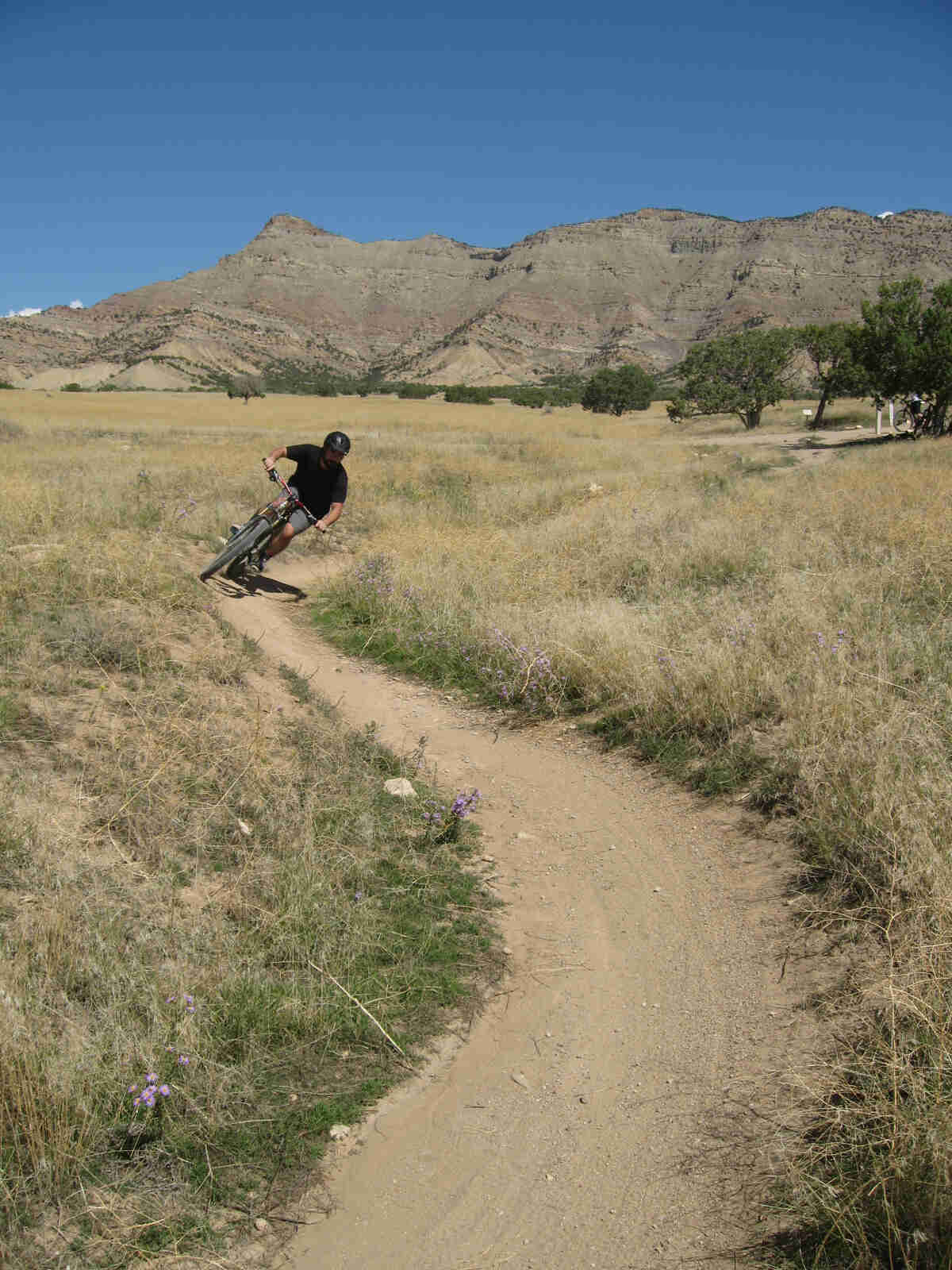 Front view of a cyclist riding a Surly bike, rounding a corner on a dirt trail, in a prairie field with mountains behind