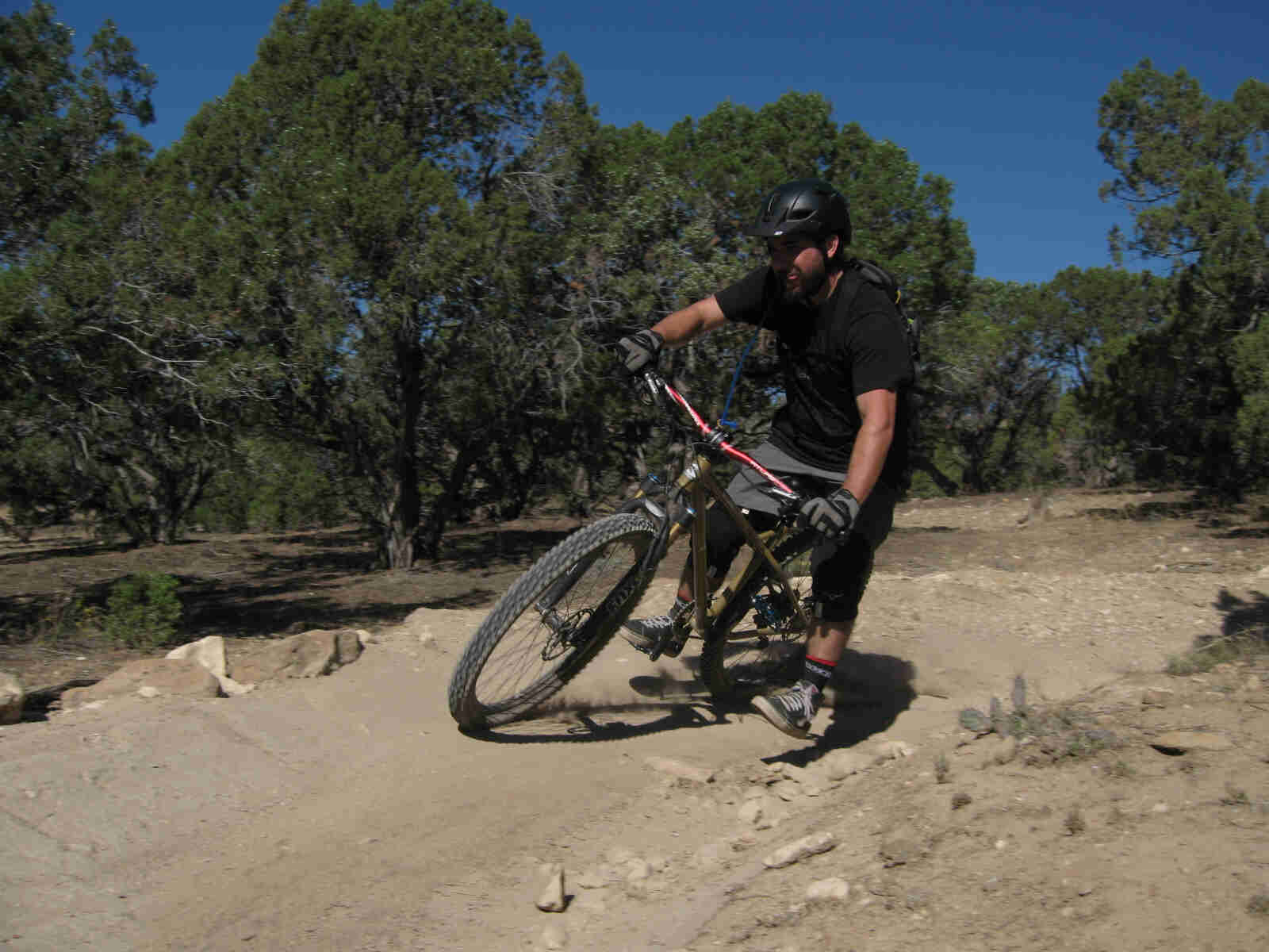 Front view of a cyclist riding a Surly Instigator bike, rounding a berm on a rocky trail, with small trees behind them