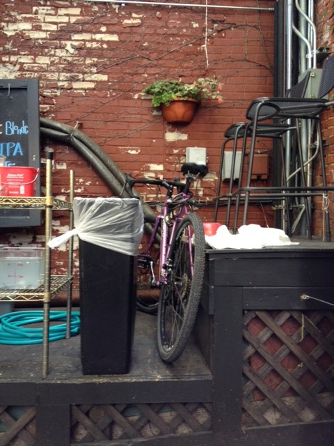 Right side view of a purple Surly bike, parked on top of a wood outdoor platform, facing a brick wall