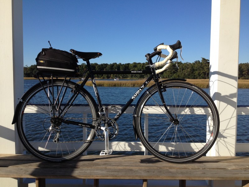 Right side view of a black Surly Cross Check bike, parked on a wood deck, above a lake with trees behind it