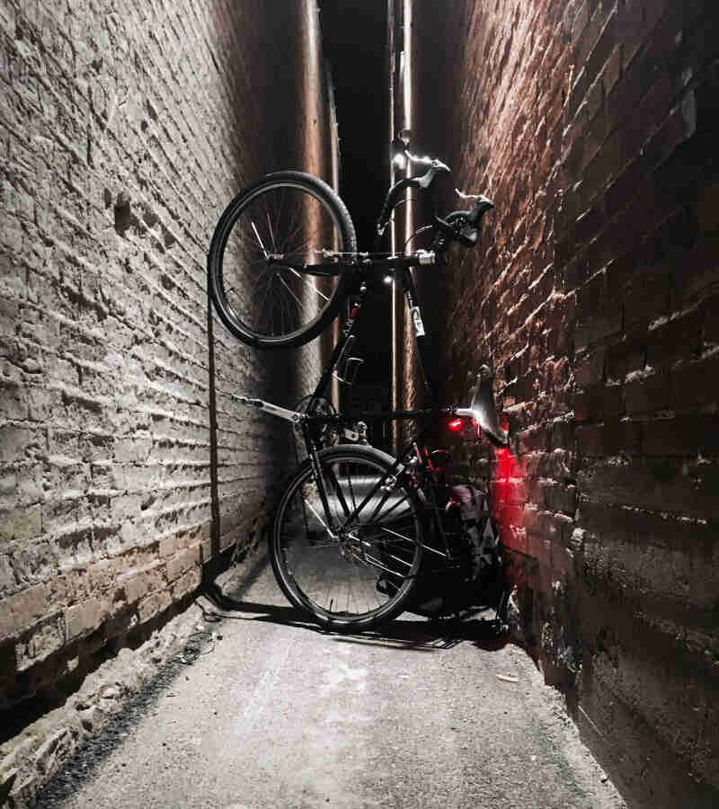 Right side view of a Surly bike, black, standing vertically between 2 brick walls, in a narrow alley at night