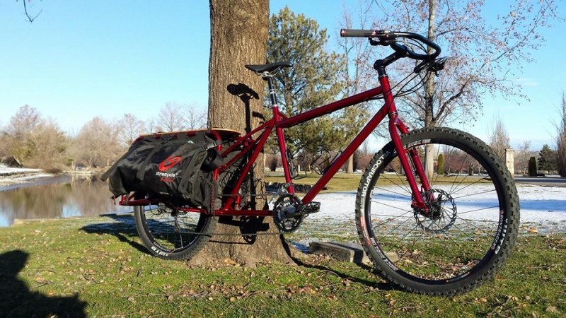 Right side view of a red Surly Big Dummy bike, parked next to a river, against a tree in a field with patches of snow