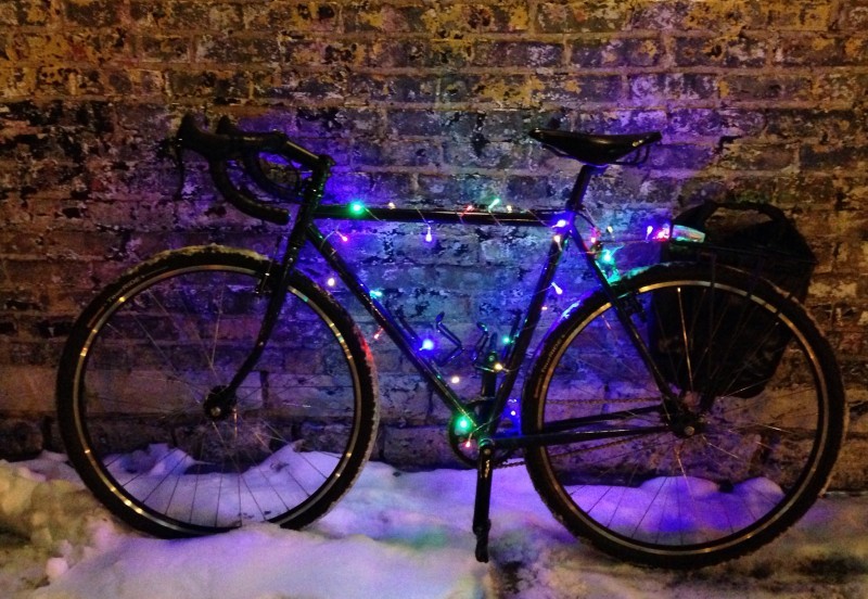 Left side view of a Surly bike with colored lights on it, parked on the snow and leaning against a brick wall at night