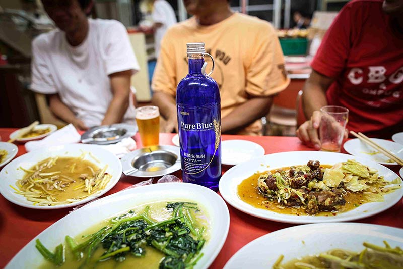 A table with plate of food on top and a bottle of Pure Blue liquor in the middle, with people sitting behind it
