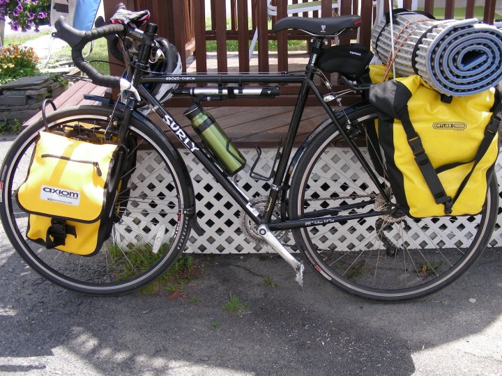 Left side view of a black Surly Cross Check bike, loaded with gear, parked on pavement against a wood deck