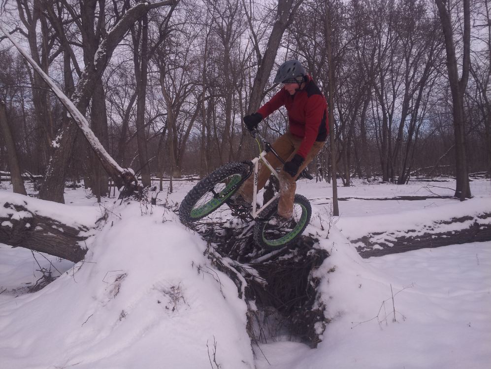 Front, right side view of a cyclist, on a white Surly Pugsley fat bike over roots of a downed tree, in the snowy woods