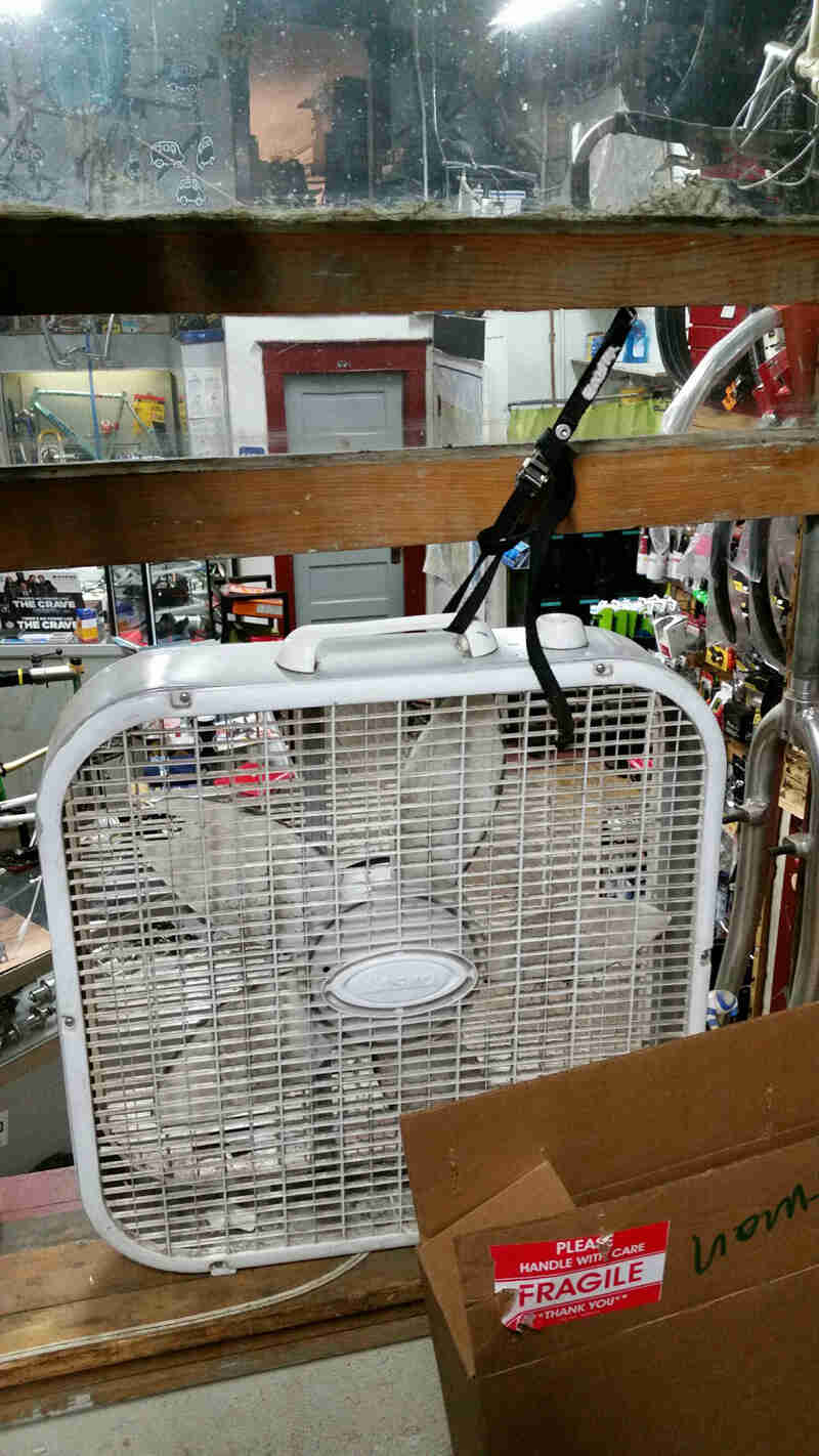 A Surly Junk strap - black - wrapped around the handle of a box fan and a wood rail