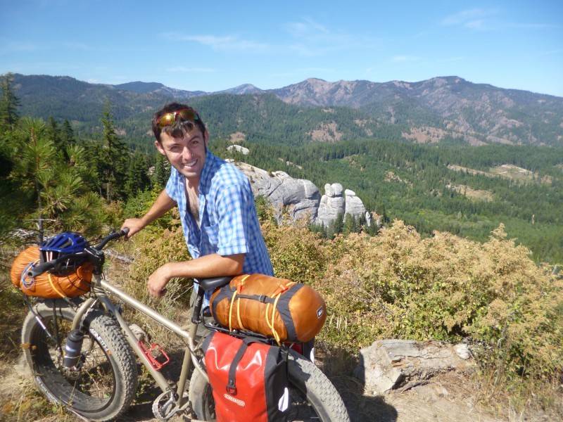 Left side view of a Surly fat bike with gear, with a cyclist on right side, on a hilltop with mountains in background
