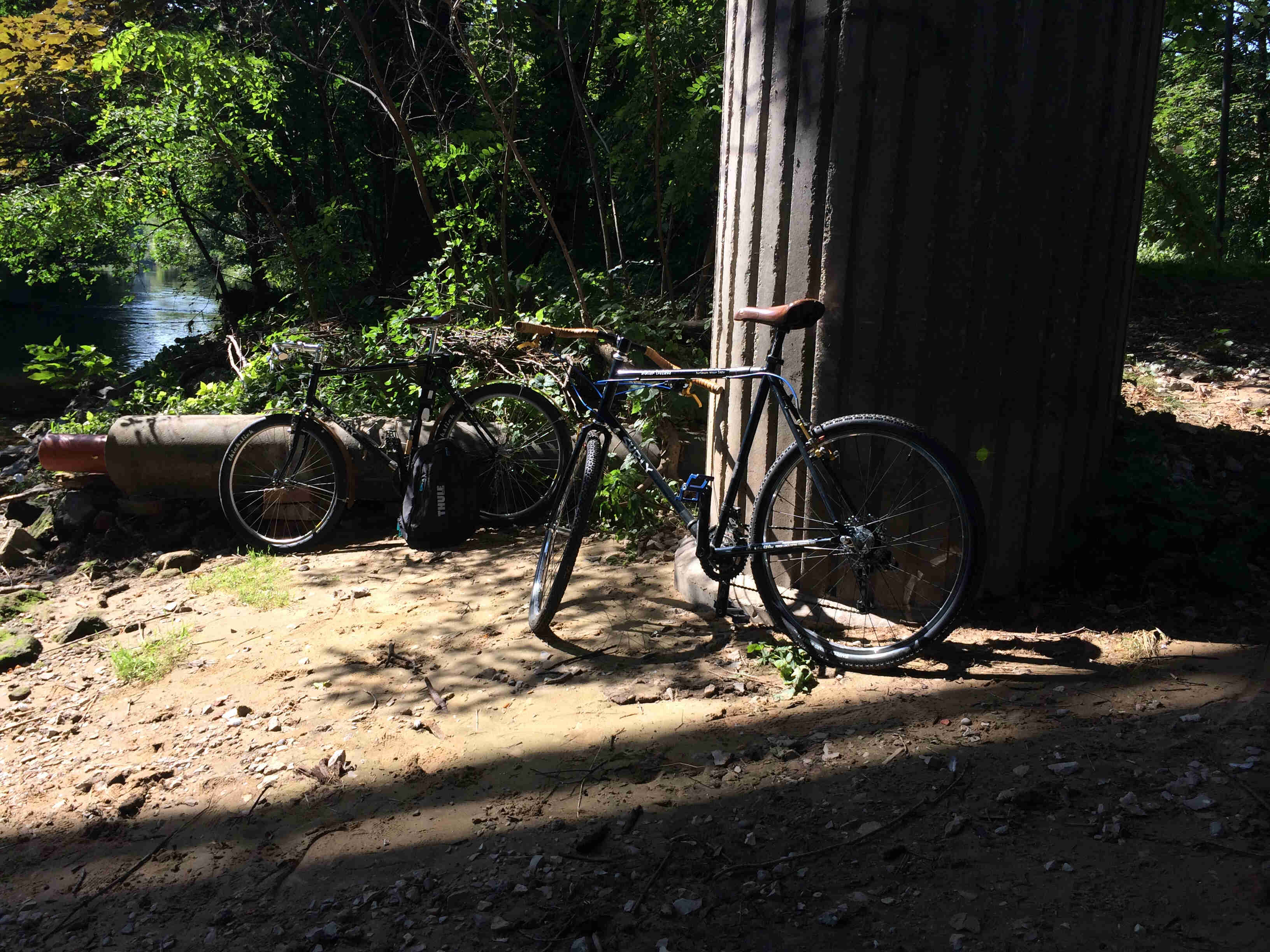 Right side view of 2 Surly bikes, parked on a flat dirt bank near a river in the woods, next to a bridge pillar