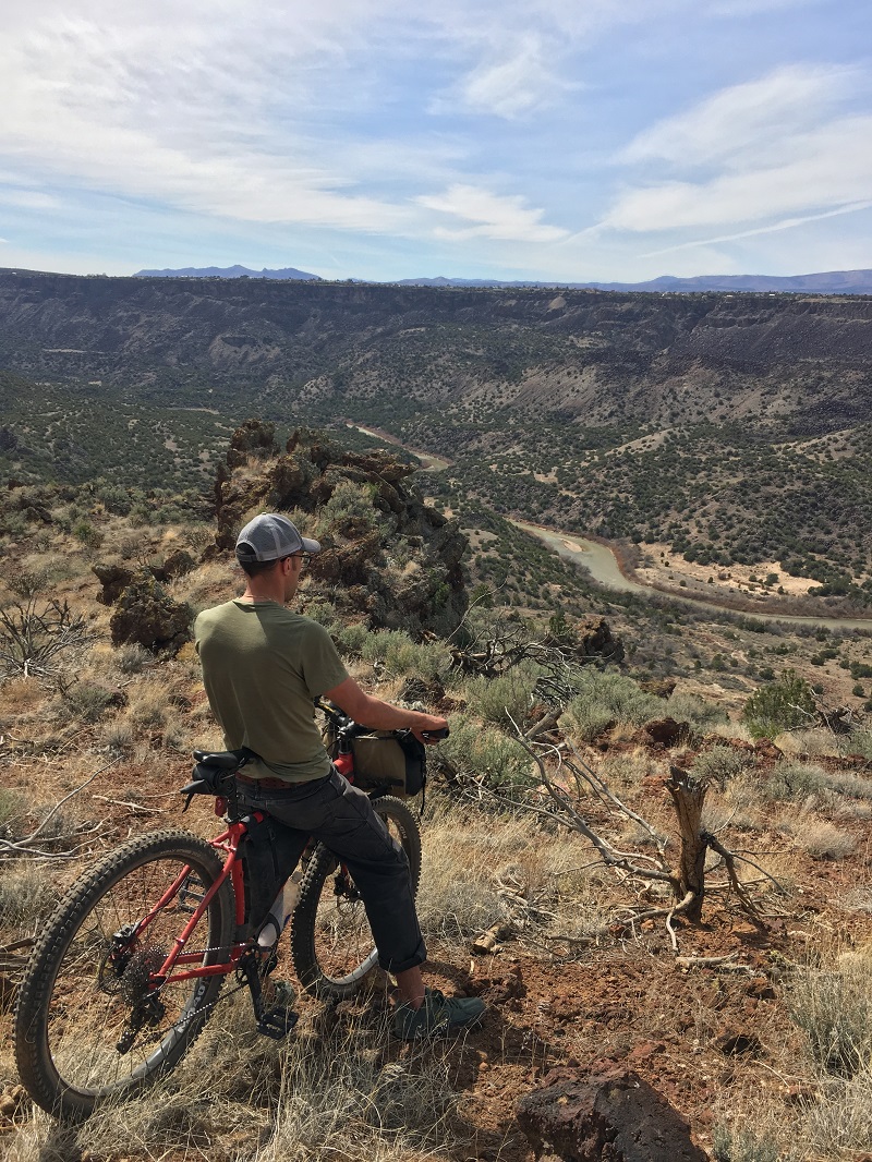 Cyclist stands with a red bike and looks down at a desert river canyon with hills, and blue sky in the background