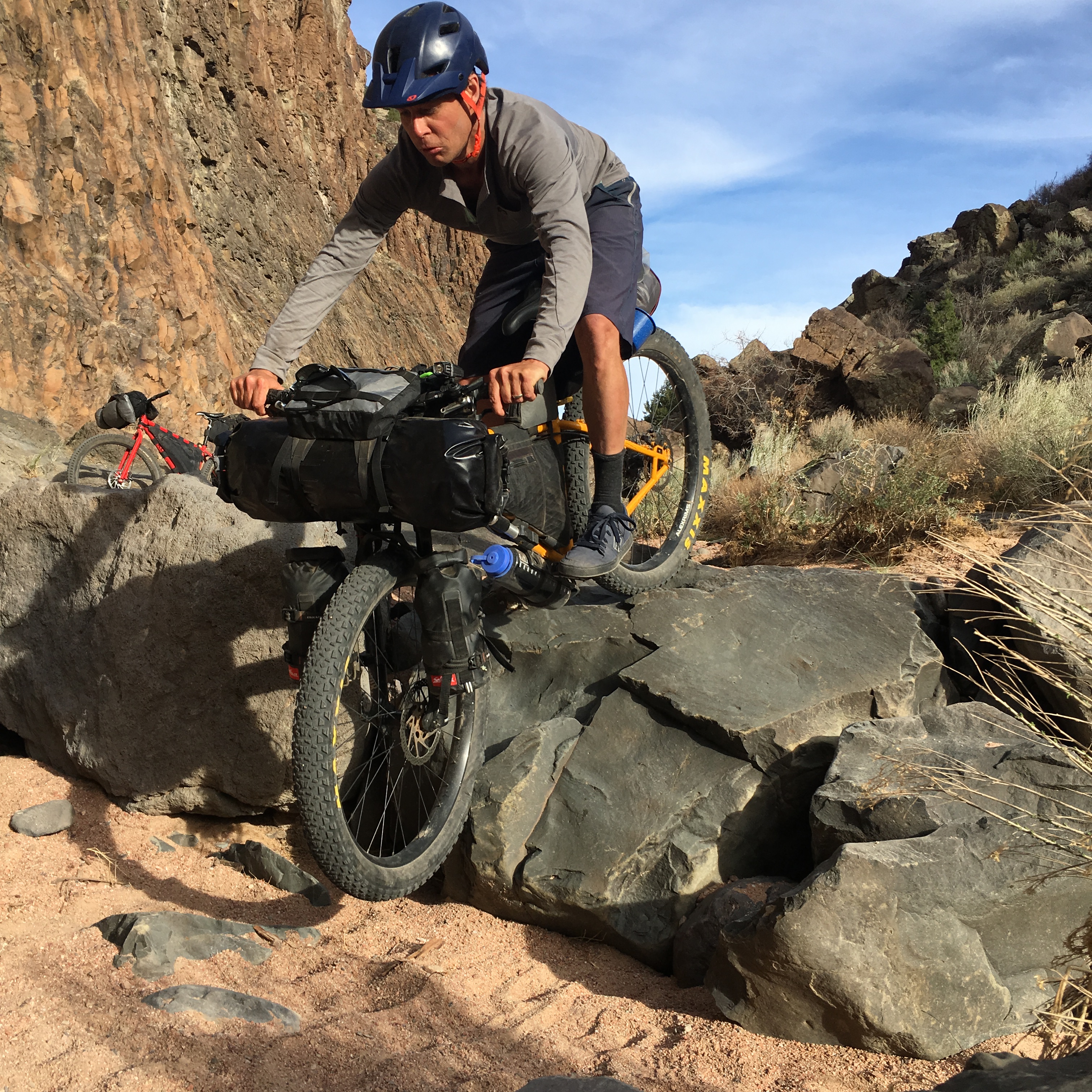 Front view of a cyclist, riding a Surly bike off of a rock in a desert canyon, with a red bike in the background