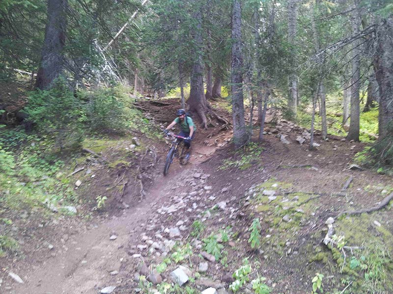 Front view of a cyclist, riding a Surly Instigator bike down a rocky dirt trail, in the forest
