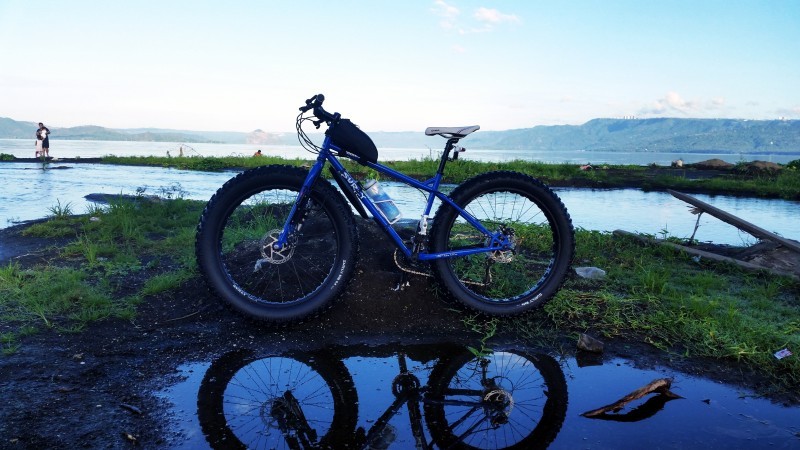 Left side view of a blue Surly bike, leaning on a grass bank behind a puddle, with water and land in the background