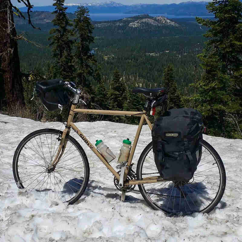 Left side view of a Surly bike, standing on a snowy hilltop, overlooking a forest valley, with mountains in the distance