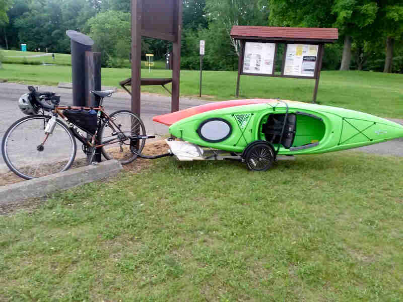 Left side view of a bike with a trailer hauling kayaks hitched behind, parked next to a lot at a park