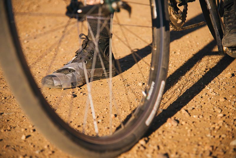 Zoomed in view of the lower half of a bike wheel in sand with a black, high top shoe standing behind