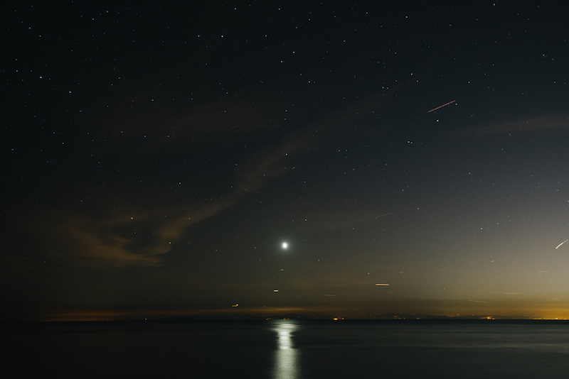 A calm body of water with stars and the moon in the sky at night