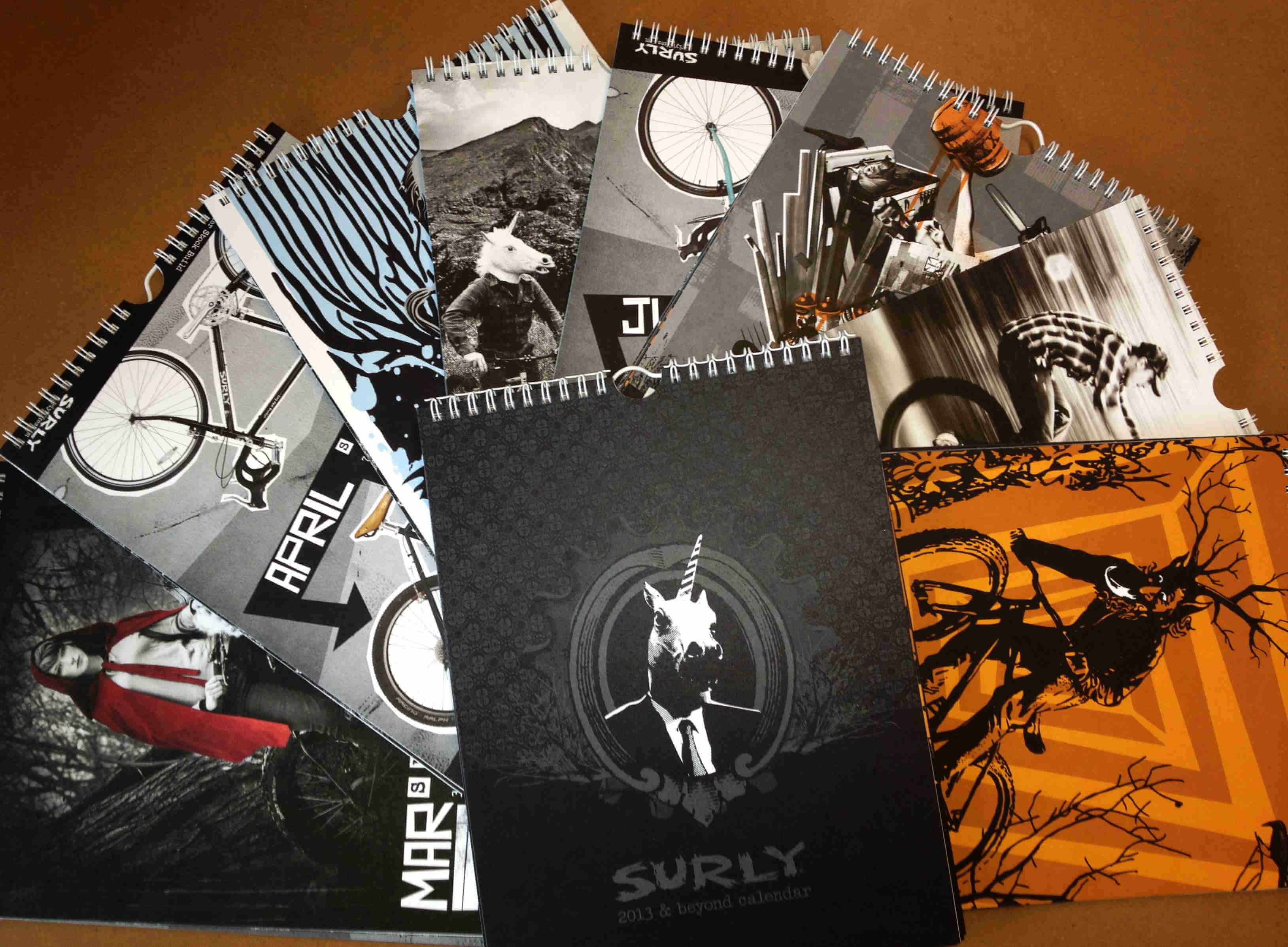 Downward view of 2013 Surly Bikes calendars, opened to different pages, laying on top of each other, on a brown surface
