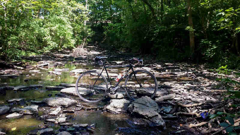 Right side view of a black Surly Cross Check bike, parked in the middle of a low, rocky stream in the woods