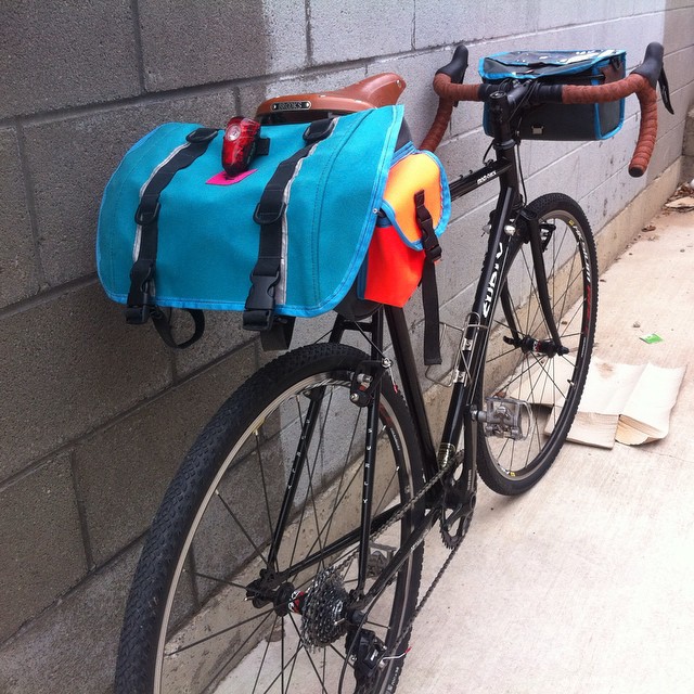 Rear, right side view of a black Surly Cross Check bike, with handlebar and seat packs, leaning on a cinder block wall