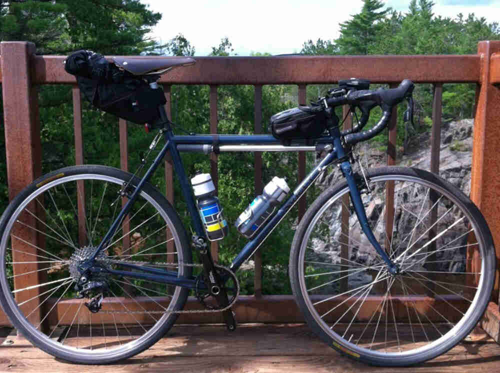 Right side view of a blue Surly Cross Check bike, parked against a rusty handrail, with trees and a cliff behind