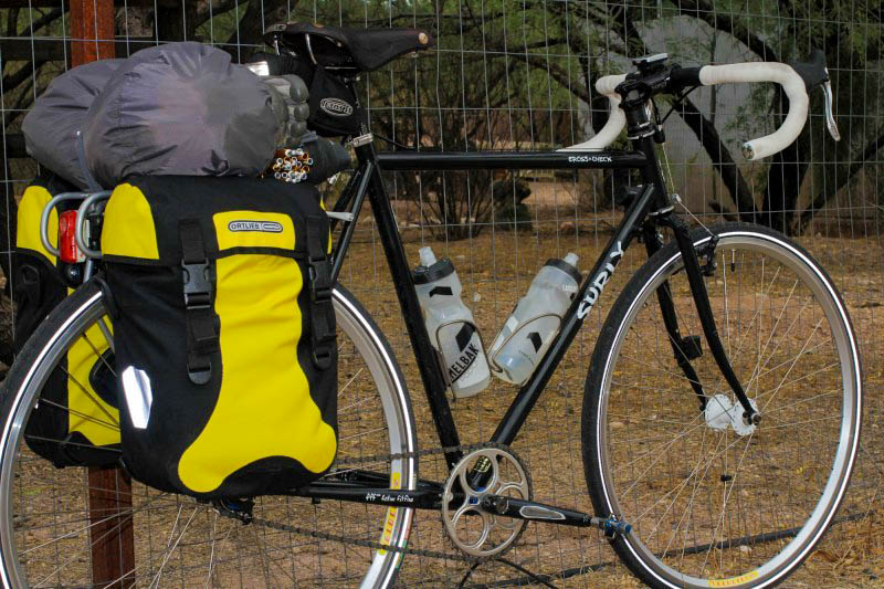 Right side view of a black Surly Cross Check bike loaded with gear on the back, parked against a tall wire fence