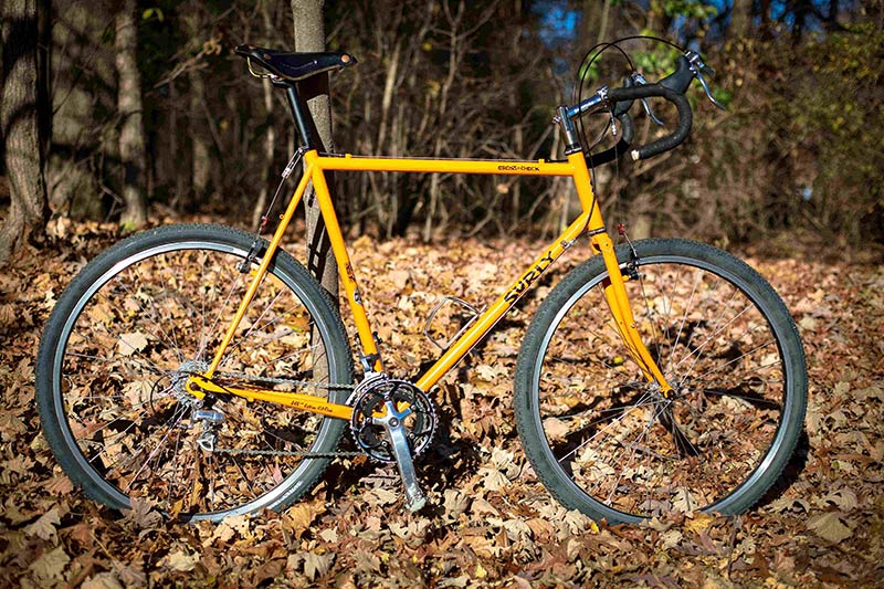 Right side view of a yellow Surly Cross Check bike, parked on leaves, with woods in the background