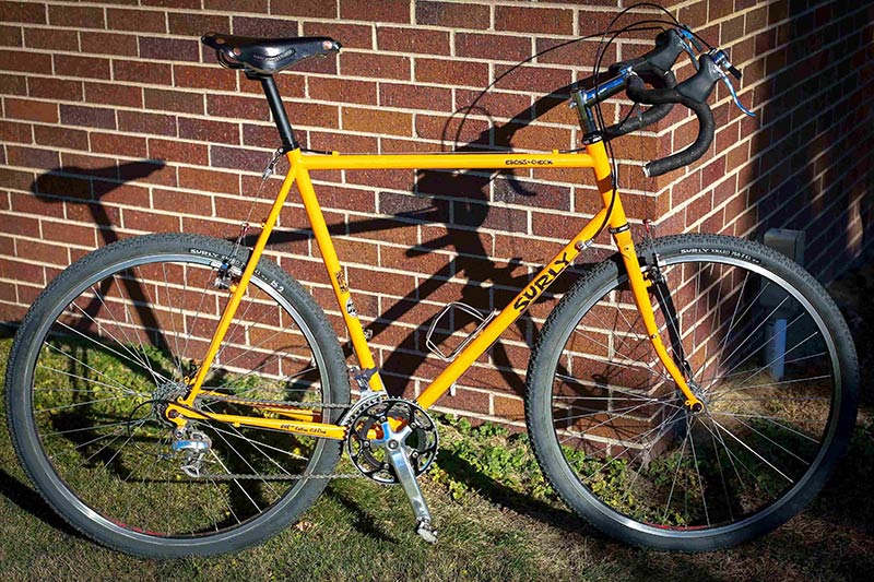 Right side view of a yellow Surly Cross Check bike, parked on grass, leaning against a brick wall