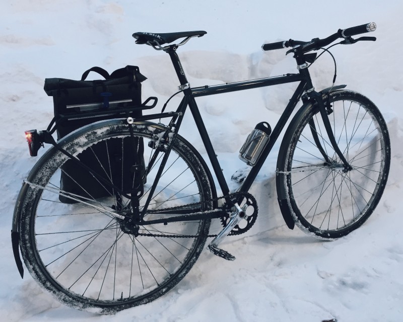 Right side view of a black Surly Cross Check bike with a left rear saddlebag, parked against a snowbank
