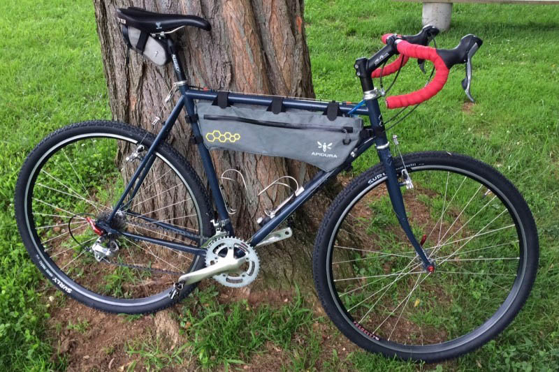 Right side view of a black Surly Cross Check bike with a frame pack, parked on grass, leaning on the base of a tree