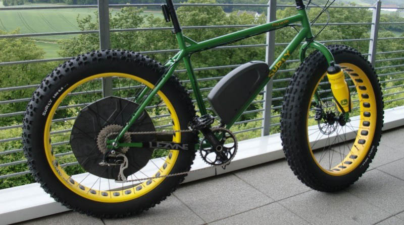 Right side view of a green and yellow Surly fat bike, parked against a railing, with trees below in the background