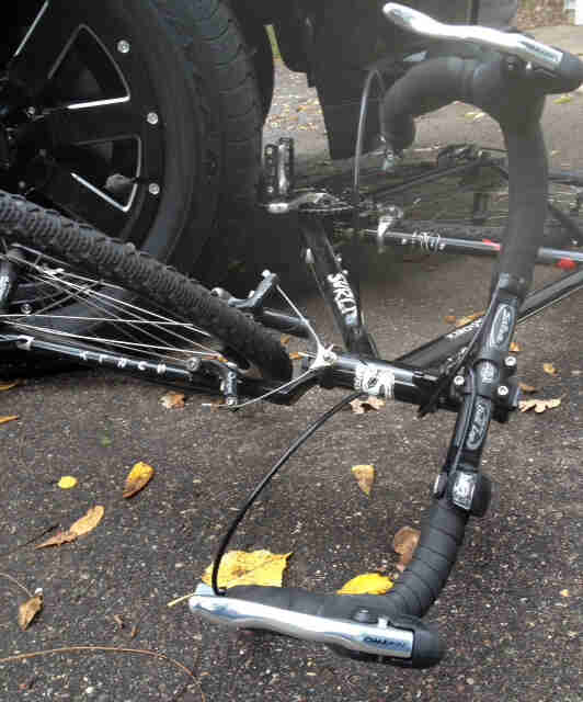 Front view of a black Surly Cross Check bike, laying on it's left side, on pavement under a car wheel