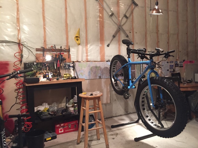 Front, right side view of a blue Surly fat bike, mounted on a stand, with the back wheel near a wall, inside a workshop