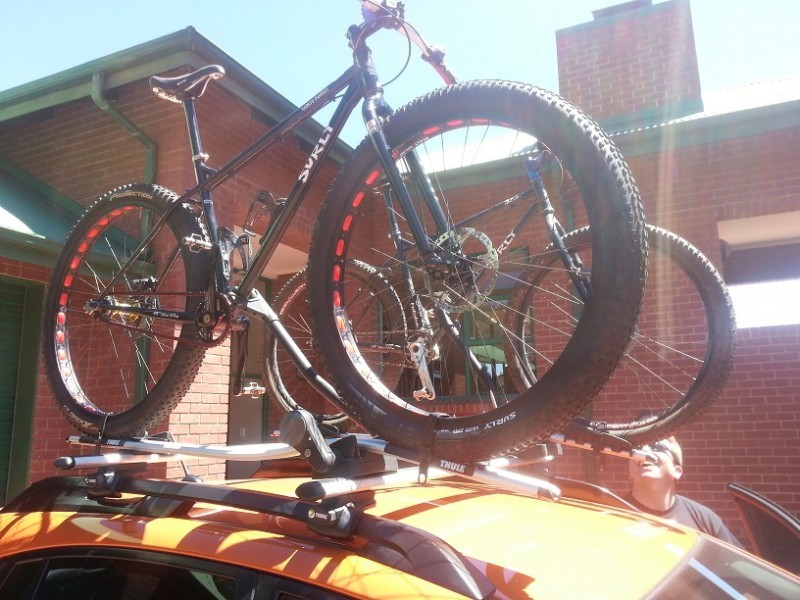 Upward, right side view of a black Surly Karate Monkey bike, mounted in a rack on a car roof, with a building behind