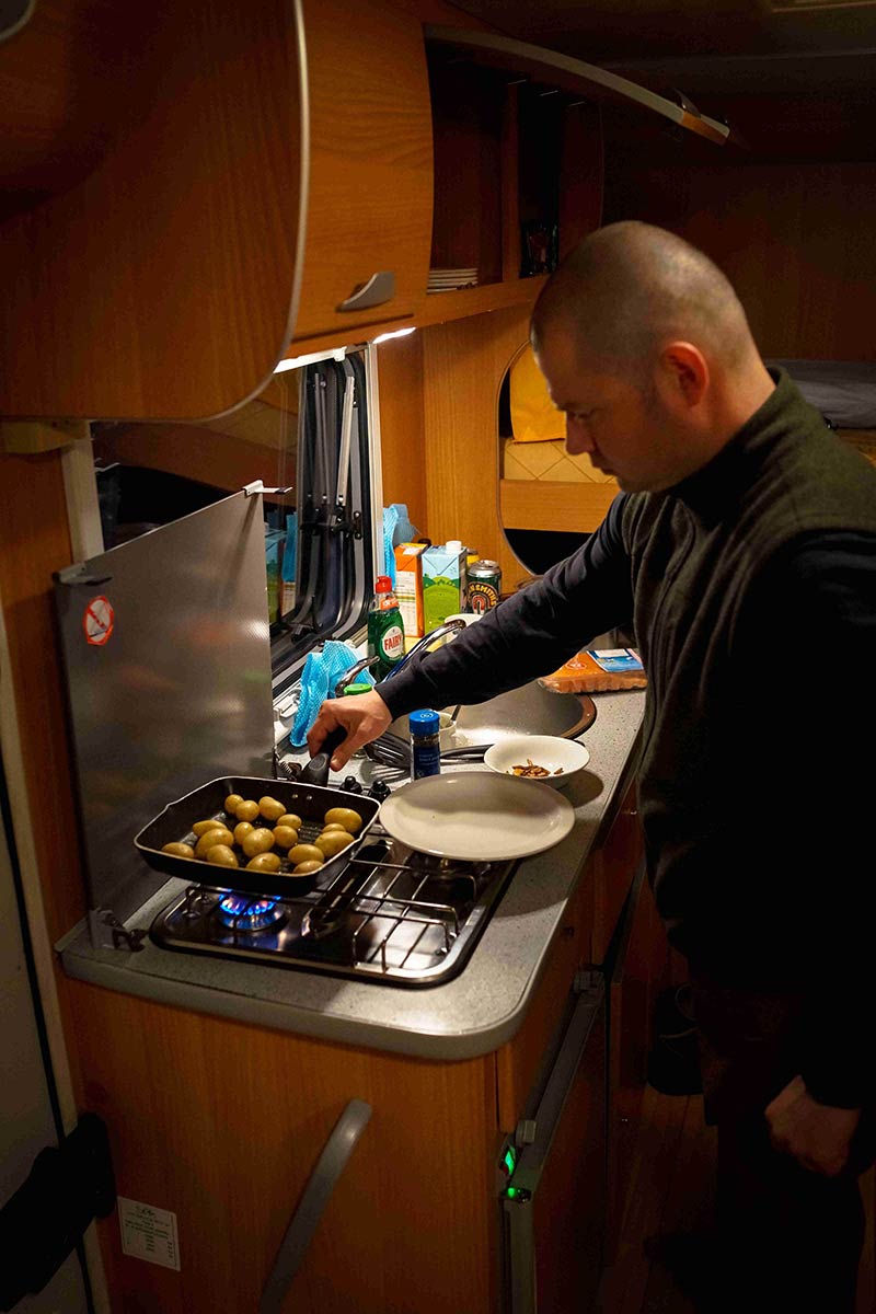 Left side view of a person, cooking a pan of small potatoes, on a stovetop in a motorhome