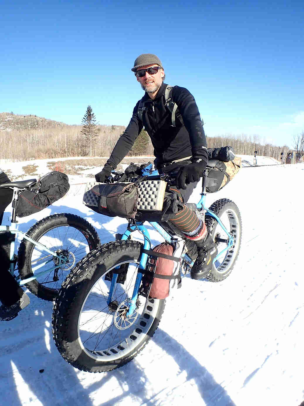 Front view of a cyclist riding a blue Surly Ice Cream Truck fat bike loaded with gear, on a snow covered field
