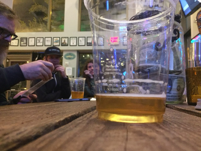 Table top view of a near empty, plastic glass of beer, on a table with people sitting around it, in an indoor room