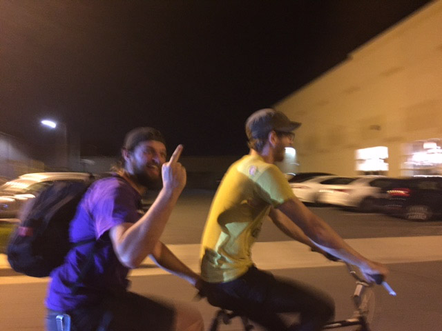 Right side view of 2 cyclists riding down a street at night, with the person in back showing a middle finger