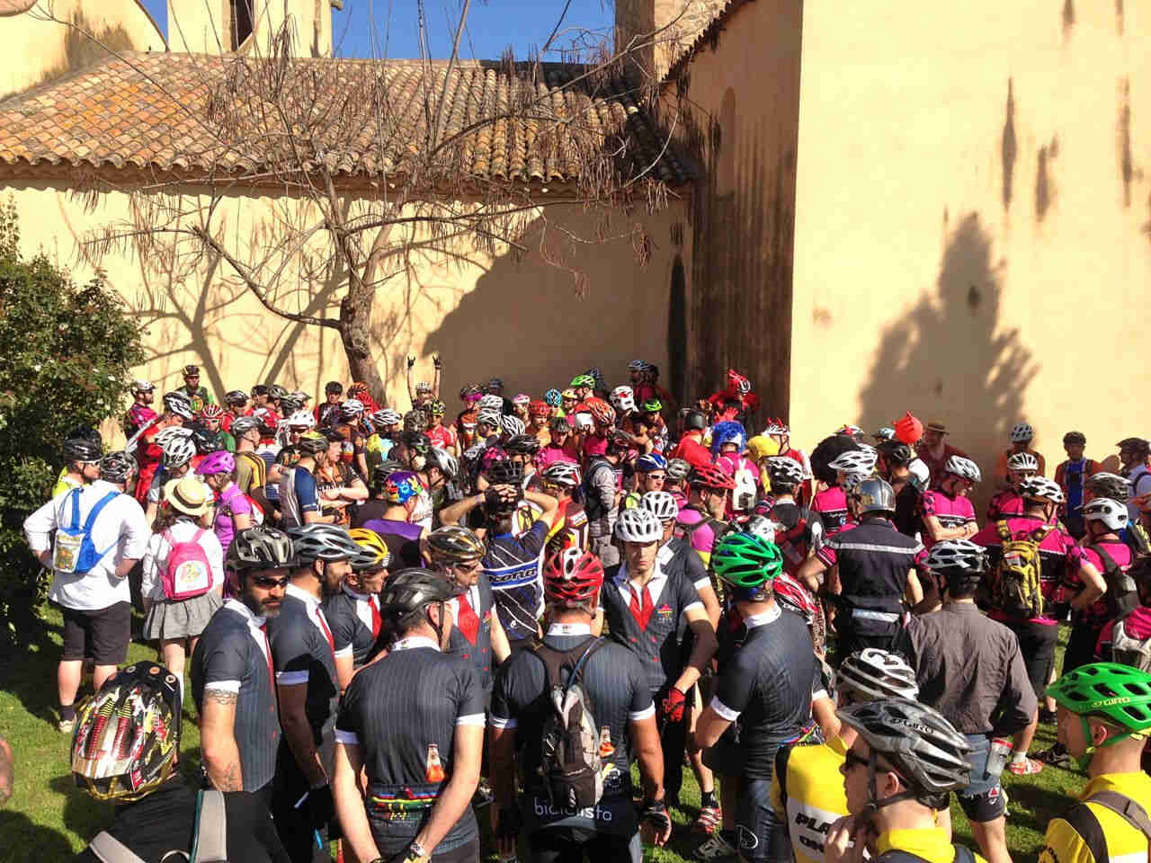 A large group of cyclists, standing closely together, outside of a church on a sunny day