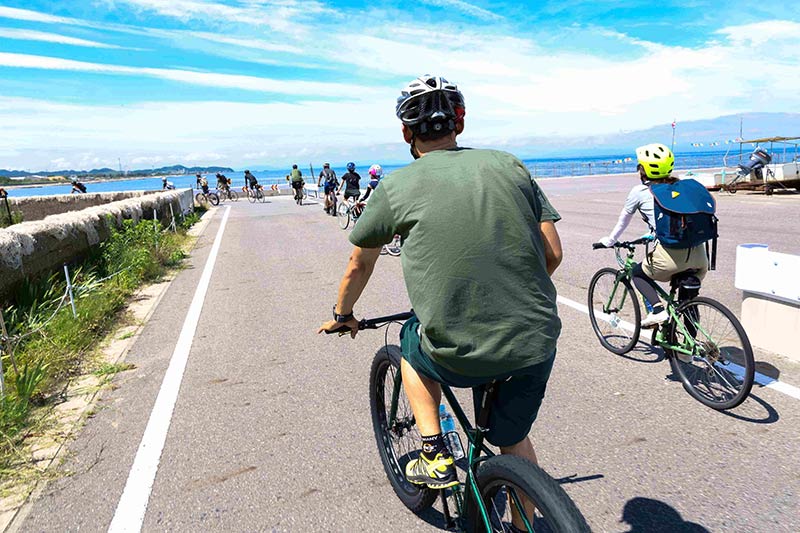Rear view of a cyclist, riding a green Surly Krampus bike on a paved road with others, with a body of water ahead