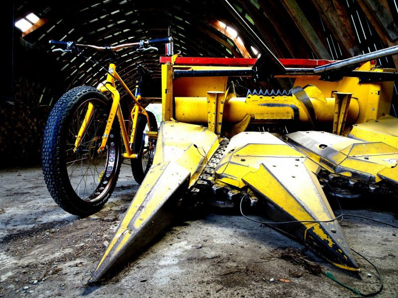 Front view of a yellow Surly Pugsley bike, leaning against a front part from a farming combine, in a barn